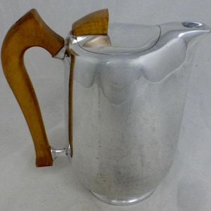 Puede incluir: Una cafetera de plata con un mango de madera. La cafetera tiene un cuerpo redondeado y un pico en el lateral. El mango es de color marrón oscuro y está unido a la cafetera con una bisagra de metal.