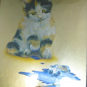 Puede incluir: Un gatito calico sentado sobre un fondo dorado con un frasco de pintura azul derramado y un pincel. El gatito tiene pelaje negro, blanco y naranja.