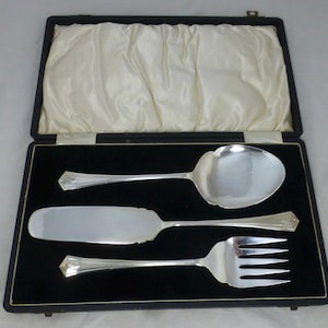 Puede incluir: Un juego de tres cubiertos de servir plateados en un estuche forrado de terciopelo negro. El juego incluye una cuchara, un cuchillo para mantequilla y un tenedor.