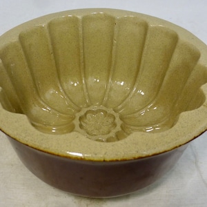 Puede incluir: Un molde de cerámica marrón y beige con un diseño interior acanalado. El molde tiene una forma redondeada con un motivo floral decorativo en el centro. El exterior es de color marrón oscuro.