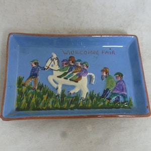 Puede incluir: Un azulejo de cerámica azul y blanco con una escena de personas montando un caballo blanco. El azulejo tiene las palabras "Widecombe Fair" escritas en él.