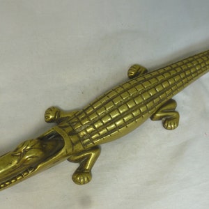 May include: A gold-colored metal alligator nutcracker with a detailed, textured body and open mouth. The alligator is lying on a white surface.