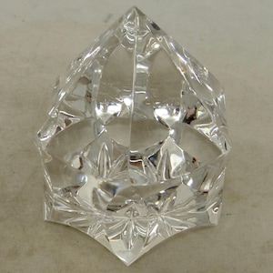 May include: A clear, faceted crystal object with a geometric design. The object has a pointed top and a starburst pattern at the base. The crystal reflects light, creating a sparkling effect. The object appears to be a decorative item.