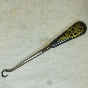 May include: A black shoe horn with a gold floral design on the handle. The handle is made of wood and the horn is made of metal.