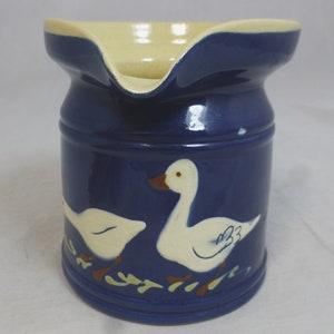May include: A blue ceramic pitcher with a white rim and two white geese painted on the side. The geese are walking on a green line with green leaves.