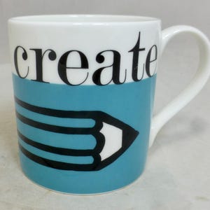 May include: White ceramic mug with a blue band and the word "create" in black letters. A black and white pencil graphic is on the blue band. The mug has a white handle.