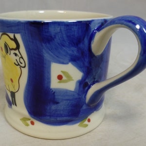 May include: A white ceramic mug with a blue and yellow hand-painted design of a sheep. The mug has a white handle and a white rim.