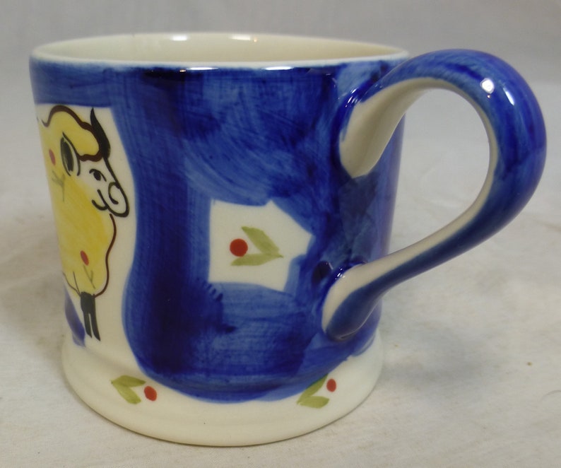 May include: A white ceramic mug with a blue and yellow hand-painted design of a sheep. The mug has a white handle and a white rim.