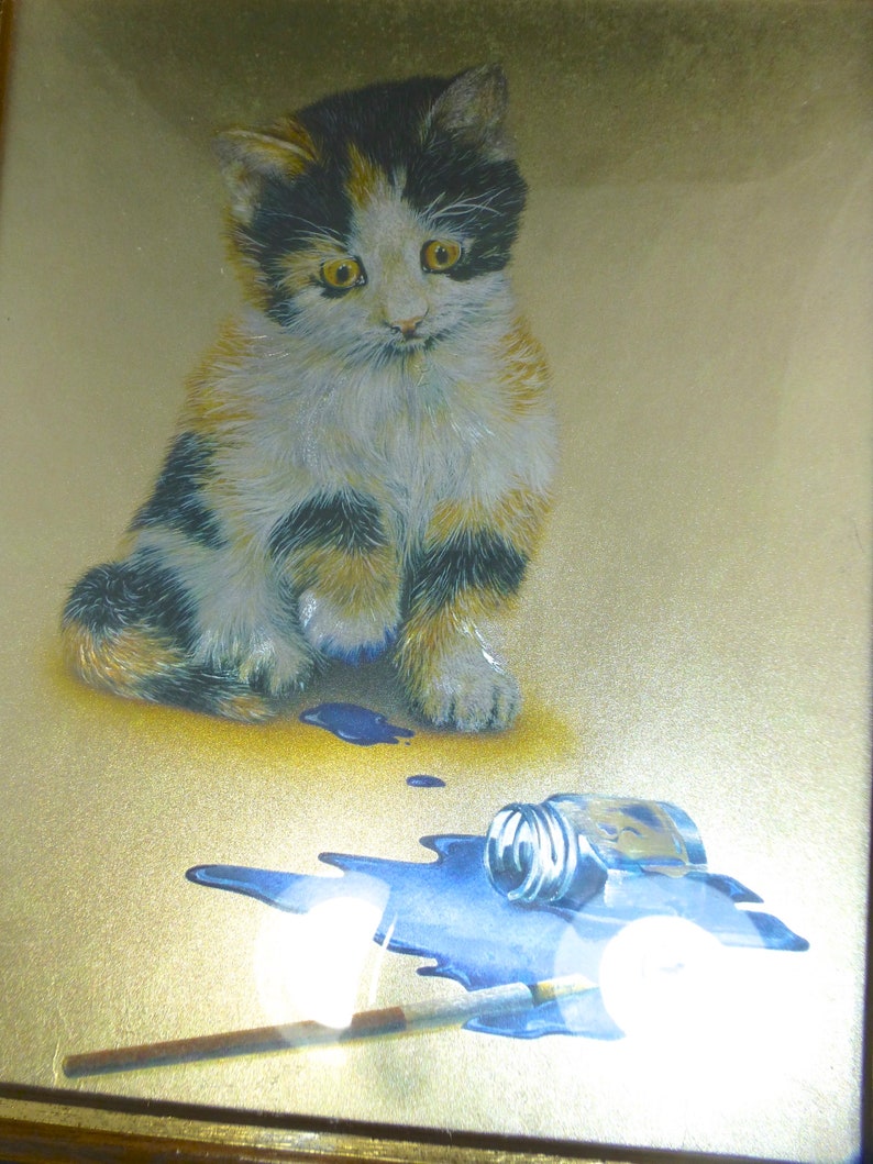 Puede incluir: Un gatito calico sentado sobre un fondo dorado con un frasco de pintura azul derramado y un pincel. El gatito tiene pelaje negro, blanco y naranja.