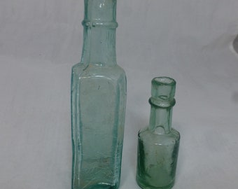 May include: Two vintage glass bottles with a light green tint. The larger bottle is rectangular with a slightly flared top. The smaller bottle is rounder with a shorter neck. Both bottles have a textured surface.