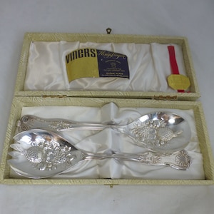 May include: A set of two silver salad servers in a cream-coloured box with a satin lining. The servers have a decorative design of fruit and flowers. The box has a label that reads "Viners Kingsly Royal Alpha Plate Sheffield England".