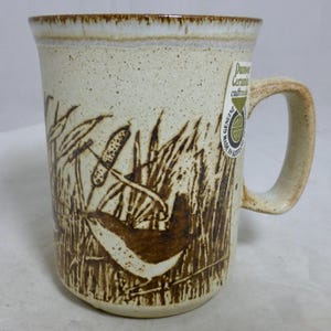 May include: A white ceramic mug with a brown design of a bird in reeds. The mug has a brown handle and a sticker that says "Genuine Ceramic Craftsmanship".