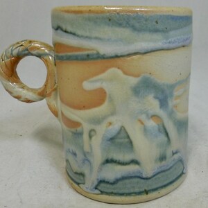 Puede incluir: Una taza de cerámica con cuerpo cilíndrico y asa en forma de cuerda trenzada. La superficie exterior muestra un efecto de esmalte en capas en tonos crema, melocotón suave y azul apagado, con gotas de esmalte blanco que caen.