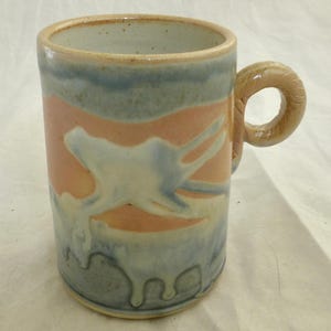 Peut inclure: Une tasse en céramique cylindrique présentant un motif d'émail aux tons pêche doux, bleu pâle et blanc. La tasse possède une anse épaisse et arrondie ainsi qu'un bord intérieur lisse et moucheté. L'émail extérieur est appliqué en couches.