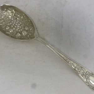 Puede incluir: Una cuchara de servir de plata con un diseño ornamentado que presenta un racimo de uvas y hojas. La cuchara tiene un mango largo y delgado con un patrón decorativo.