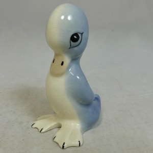 Puede incluir: Figura de pato de cerámica en azul claro y crema. El pato tiene una cabeza grande, un pico pequeño y ojos negros. Sus patas son negras y está sentado. Un artículo decorativo.