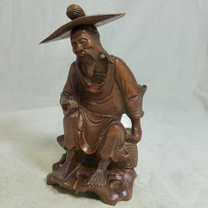 May include: A detailed wooden sculpture of an elderly man wearing a wide-brimmed hat. The figure is seated, with a long beard and traditional clothing. The sculpture is carved from a dark wood and rests on a carved base, showcasing intricate details.