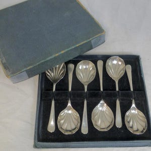 May include: A set of six silver-toned spoons with shell-shaped bowls and smooth handles, arranged in a dark blue presentation box with a black velvet interior. The spoons feature a fluted design on the bowl, and the box has a matching lid.