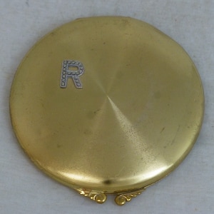 Puede incluir: Un espejo compacto de oro con una letra "R" enjoyada en la tapa.