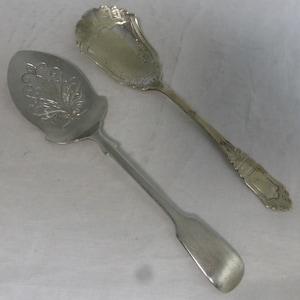 Può includere: Due cucchiai da portata in argento con manici decorati. Il cucchiaio a sinistra ha un motivo floreale decorativo sulla ciotola. Il cucchiaio a destra ha un design più elaborato sul manico.