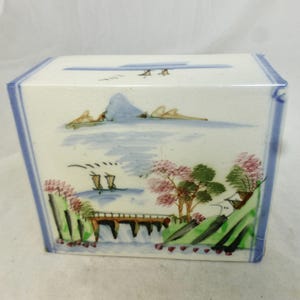May include: A white ceramic box with a hand-painted landscape scene. The scene includes a mountain, water, trees with pink blossoms, and a bridge. The box has blue trim and a lid with a similar landscape design.