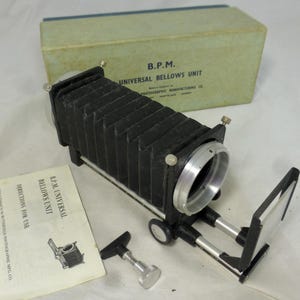 May include: A vintage B.P.M. Universal Bellows Unit camera attachment with a black bellows, silver lens ring, and a light green box. Includes a manual and a silver-colored adjustment tool. The box reads "B.P.M. UNIVERSAL BELLOWS UNIT".