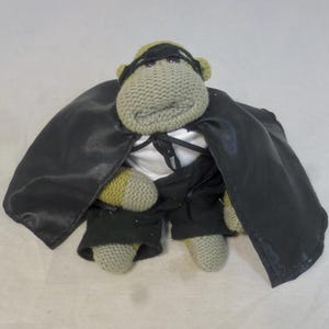 May include: A knitted monkey toy dressed as a superhero, wearing a black cape and mask, and a tuxedo. The monkey is grey with yellow details. The cape is made of a shiny black material.