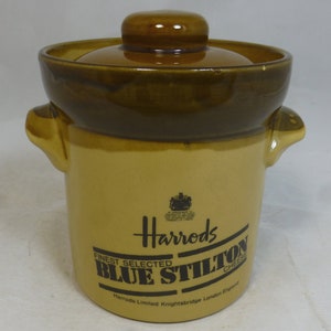 May include: A brown ceramic jar with a lid, featuring the Harrods logo and the text "Finest Selected Blue Stilton Cheese". The jar has two handles and is decorated with a brown glaze.