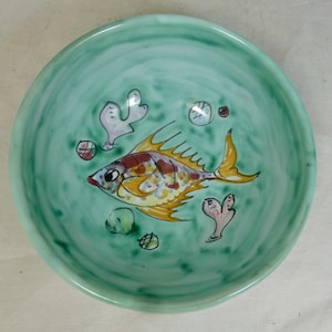 Könnte beinhalten: Eine handbemalte Keramikschale mit türkisfarbenem Hintergrund. Die Schale zeigt einen bunten Fisch mit gelben, braunen und roten Schuppen. Dekorative Meerespflanzen und Blasen sind ebenfalls auf der Schale gemalt.