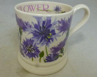 Può includere: Una tazza in ceramica bianca con un motivo floreale blu e viola. La tazza ha un manico e la parola "FLOWER" è stampata sul lato.