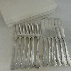 May include: A set of silver-colored forks and knives arranged on a light-colored surface. The forks have four tines and a geometric handle design. The knives have a curved blade and a similar handle design. A white box is in the background.