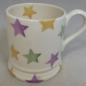 May include: White ceramic mug decorated with colourful stars in yellow, green, and purple. The mug has a curved handle and a slightly flared base. A simple, yet cheerful design for everyday use.