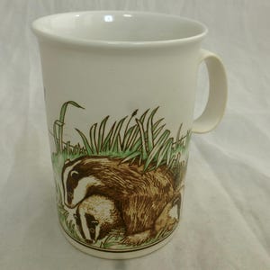 May include: A white ceramic mug featuring a detailed illustration of two badgers in a grassy field. The badgers are depicted with their characteristic black and white striped faces and brown fur, surrounded by green blades of grass.