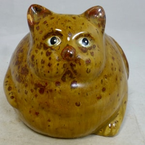 Könnte beinhalten: Eine Keramikfigur eines braunen und braunen Katers mit schwarzen Augen. Die Katze sitzt mit den Beinen unter dem Körper. Die Katze hat ein geflecktes Muster.