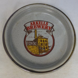 Könnte beinhalten: Ein grauer Keramik-Bieruntersetzer mit einem roten und gelben Logo, auf dem "Arkel's Brewery" steht. Das Logo zeigt ein Gebäude mit einem Schornstein und einer Backsteinmauer.