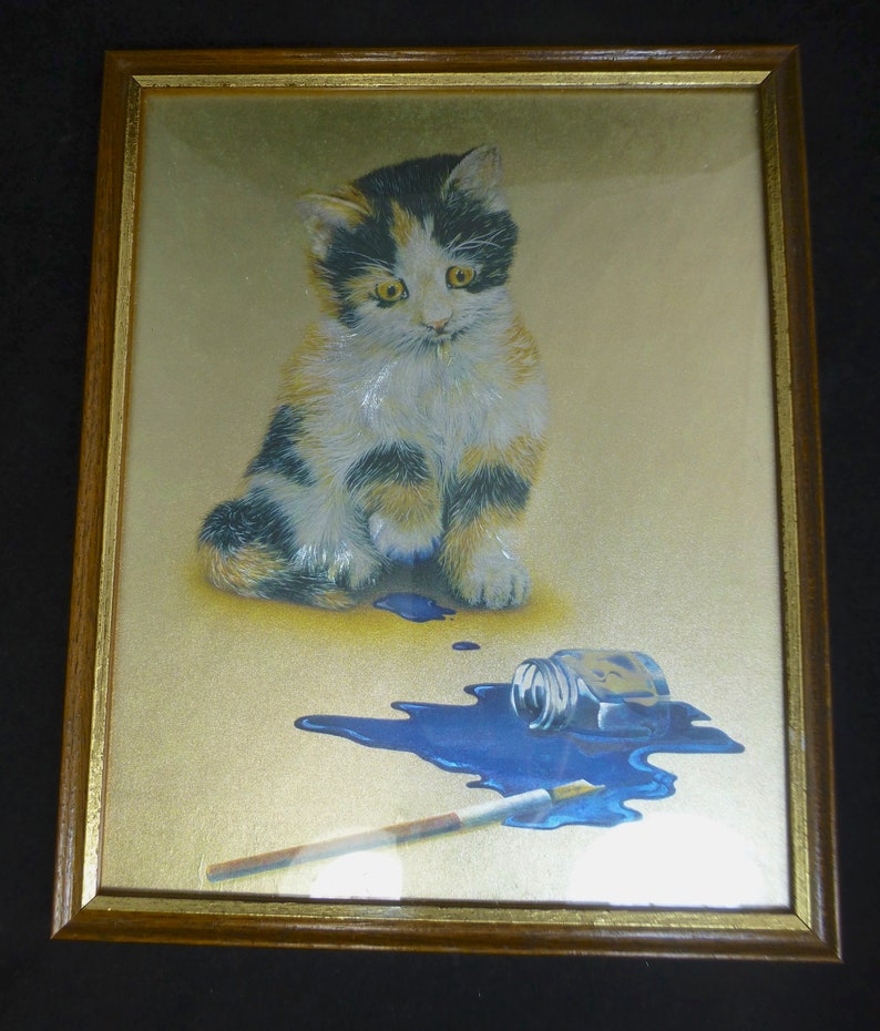 Puede incluir: Una impresi&oacute;n enmarcada de un gatito calico sentado sobre un fondo dorado con un frasco de tinta azul derramado y un pincel.