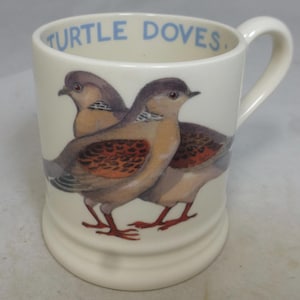 May include: Cream-colored ceramic mug featuring a detailed illustration of two turtle doves. The birds have brown, tan, and orange plumage. The words "TURTLE DOVES" are printed in blue around the rim of the mug. The mug has a white handle.