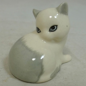 May include: A ceramic cat figurine in a seated pose. The cat is primarily white with gray markings on its back and tail. It has detailed facial features, including blue eyes and gray ears. The figurine is glossy and appears to be a decorative item.