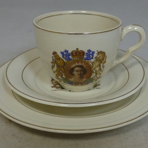 May include: A white teacup and saucer set with a gold rim. The cup and saucer feature a colorful image of Queen Elizabeth II's coronation in 1953.