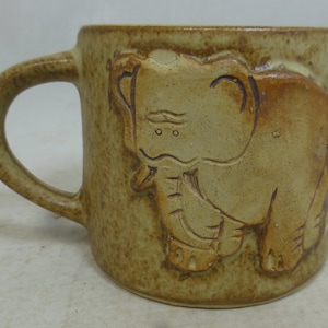 May include: A ceramic mug with a brown and tan glaze features a raised design of an elephant. The elephant is facing right and has a textured surface.