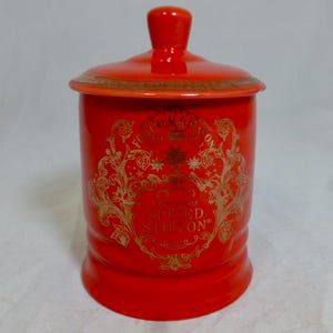 Könnte beinhalten: Ein leuchtend roter Keramikbehälter mit passendem Deckel, verziert mit goldenen Ornamenten und dem Schriftzug Fortnum & Mason sowie Christmas Spiced Stilton. Das zylindrische Gefäß hat eine glatte Oberfläche und einen Knauf am Deckel.