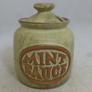 May include: A ceramic jar with a lid, in a light green color with brown speckles. The jar has a brown oval label with the words "MINT SAUCE" in brown lettering.