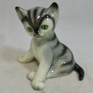 May include: A ceramic cat figurine, seated with grey and black stripes. The cat has bright green eyes and a glossy finish. This decorative item is likely for display.