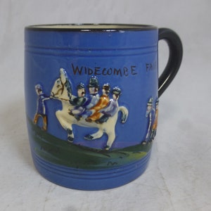 Puede incluir: Taza de cerámica azul con asa negra. La taza presenta una imagen en relieve de un caballo con jinetes y el texto "Widecombe Fair".