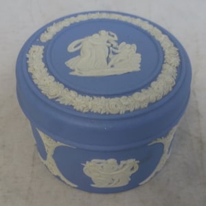 May include: A blue and white ceramic trinket box with a raised design of a classical scene. The lid features a white floral border.
