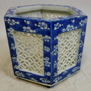 Könnte beinhalten: Ein sechseckiger Keramik-Blumentopf mit blauem und weißem Blumendesign. Der Pflanztopf hat ein gitterartiges Ausschnittdesign an den Seiten und ist mit einem blauen Rand versehen. Der Innenraum ist weiß.