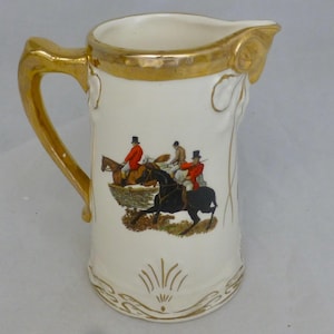 Puede incluir: Jarra de cerámica blanca con borde y asa dorados. La jarra presenta una escena de caza con tres jinetes a caballo.