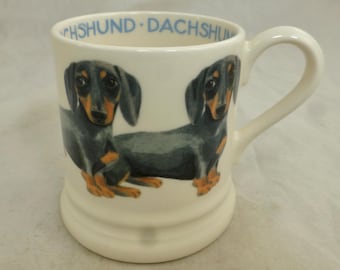 May include: White ceramic mug featuring a black and brown dachshund illustration. The mug has a blue inscription that reads "Dachshund - Dachshund".