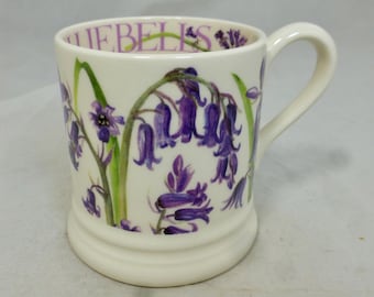 Può includere: Una tazza in ceramica bianca con un design a forma di campanula. La tazza ha un manico e le parole "Bluebells" sono stampate sul lato.