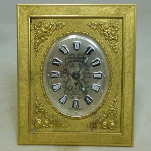 May include: A gold-coloured alarm clock with a silver face and Roman numeral markings. The clock is framed in a decorative gold frame with ornate details.